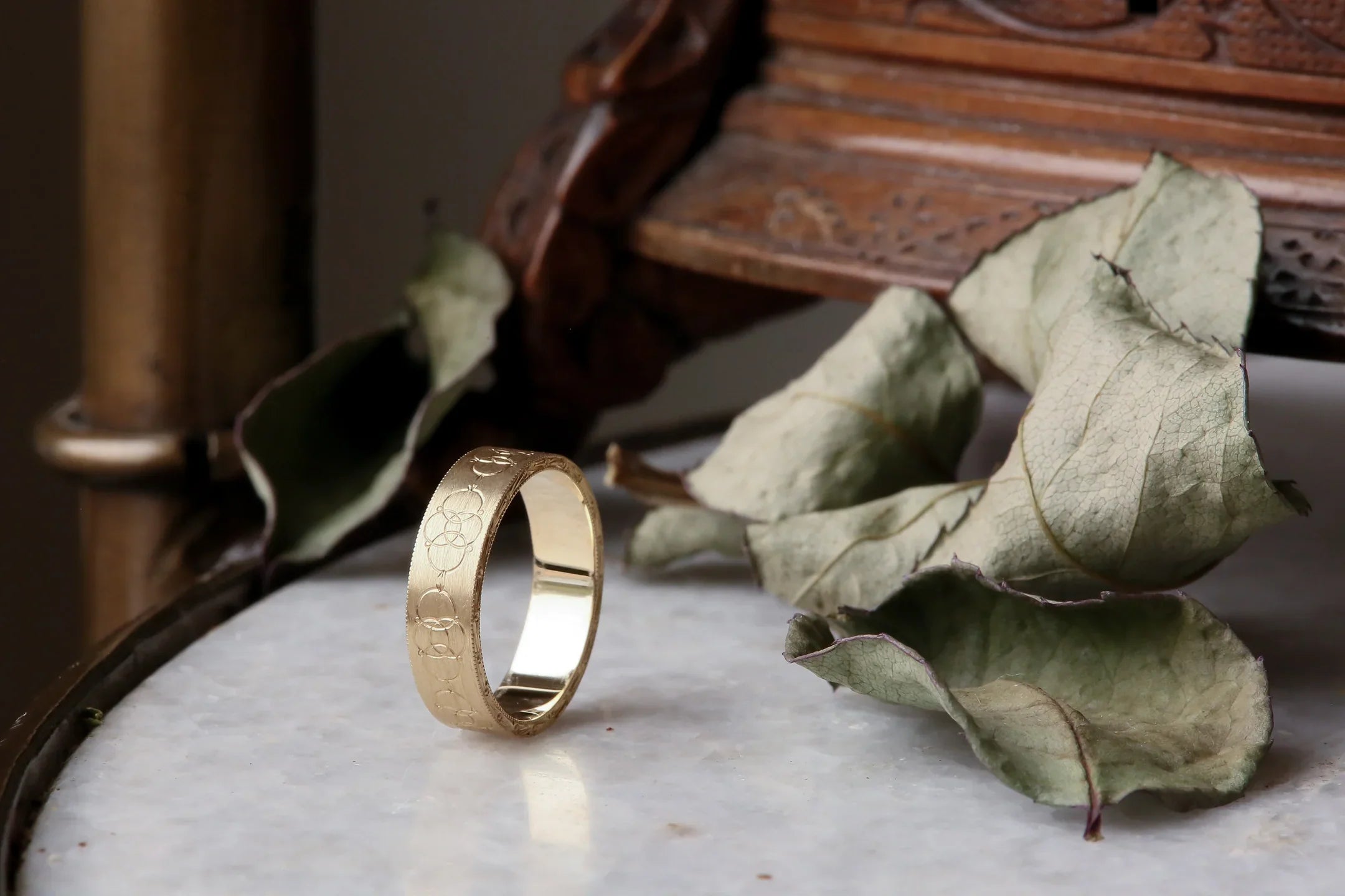 A golden ring designed and goldsmithed by The North Way Studio sits balanced on a white marble table with dark academia props scattered nearby.