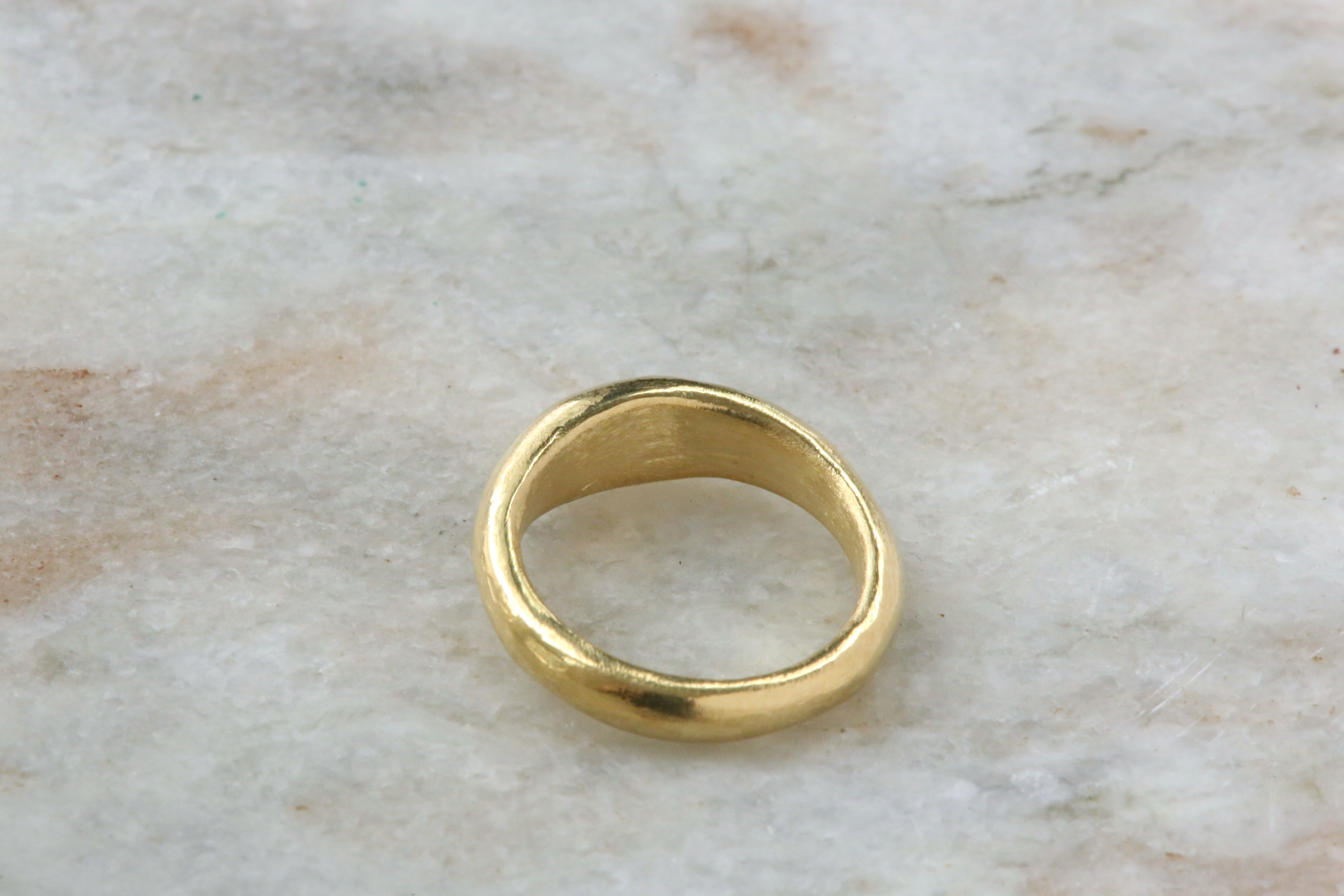 A simple gold band ring with a slightly tapered, organic shape, resting on a light beige marble surface.