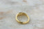 A simple gold band ring with a slightly tapered, organic shape, resting on a light beige marble surface.