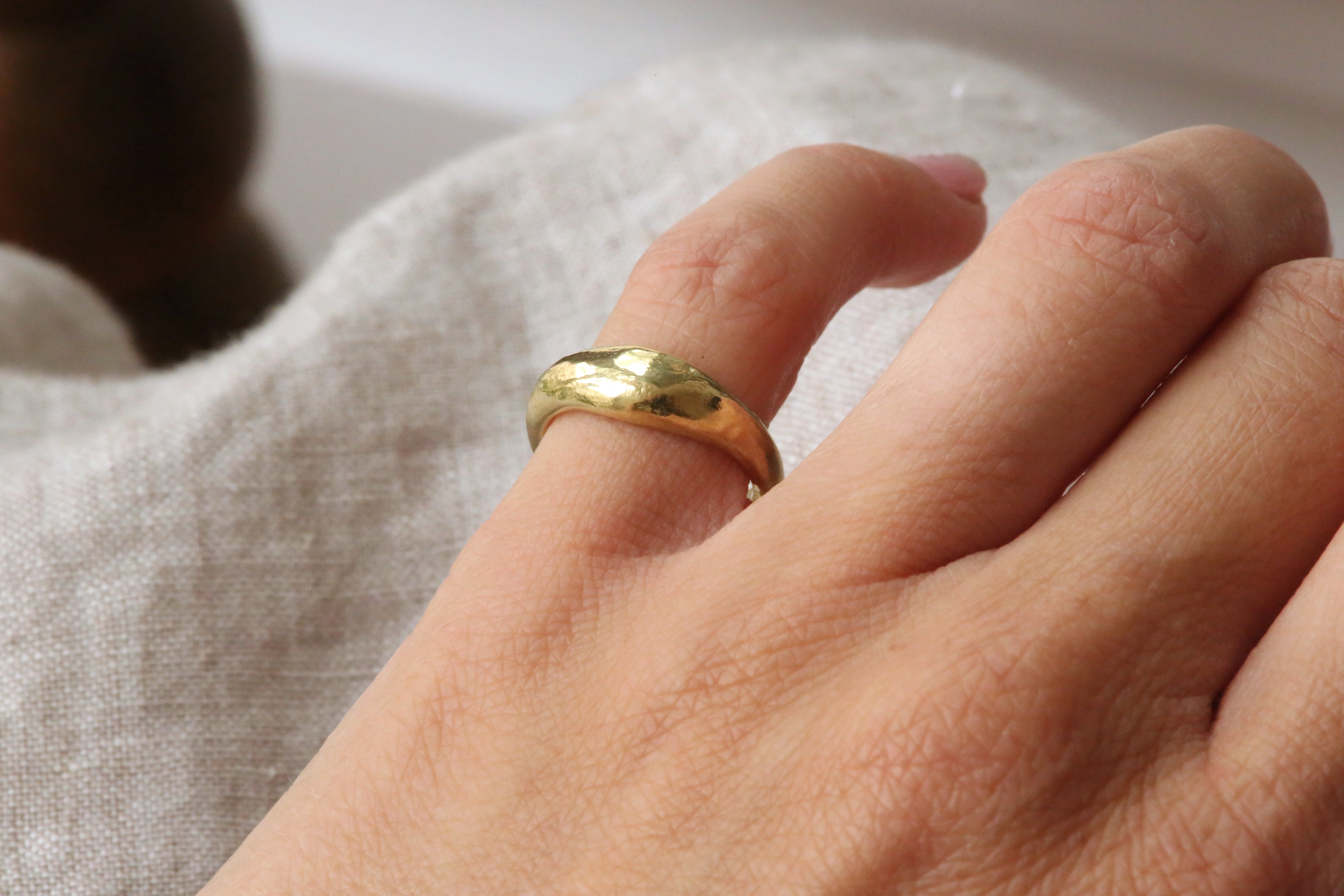 A close-up of a hand wearing a simple, polished gold band ring against a soft, light beige textured fabric background.