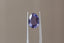 A uniquely shaped, lush portrait-cut sapphire in a regal purple hue. This sapphire is available for purchase alongside an heirloom setting at The North Way Studio. The sapphire is shown held up by tweezers.