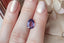 A uniquely shaped, lush portrait-cut sapphire in a regal purple hue. This sapphire is available for purchase alongside an heirloom setting at The North Way Studio. The sapphire is shown held resting on a woman's fingers for size reference.