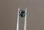 A natural round green teal sapphire available to add as a center stone for your heirloom setting at The North Way Studio. Shifting hues of teals, blues, and greens. The sapphire is shown held up by tweezers.