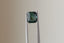 A natural emerald cut teal sapphire available to add as a center stone for your heirloom setting at The North Way Studio. A deeper forest teal hue. The sapphire is shown held up with tweezers.