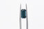 A natural elongated emerald cut teal Montana sapphire available to add as a center stone for your heirloom setting at The North Way Studio. Wisps of yellow and blue dance within this deep teal sapphire. The stone is shown held up by tweezers on a white background.