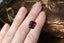 A natural emerald cut berry garnet available to add as a center stone for your heirloom setting at The North Way Studio. A lush berry wine hue worthy of royalty. The garnet is shown resting on a woman's hand amongst dried flowers.