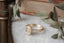 A diamond ring designed and crafted by The North Way Studio in collaboration with Claire Pettibone and cast in reclaimed gold is shown atop marble with a weathered book and dried leaves in the background.