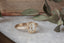 A diamond ring designed and crafted by The North Way Studio in collaboration with Claire Pettibone and cast in reclaimed gold is shown sitting atop marble with a weathered book and dried leaves in the background.