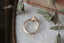 A diamond ring designed and crafted by The North Way Studio in collaboration with Claire Pettibone and cast in reclaimed gold is shown in an overhead view sitting atop marble beside a weathered book and dried leaves.