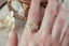 Natural diamond marquise leaves and a graduated halo are woven about an antique old European cut diamond in the softest shade of morning sun in this engagement ring by The North Way Studio and Claire Pettibone. This 18 karat yellow gold ring is shown worn on the index finger of a hand resting on an antique jewelry box.