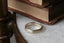 The Elder King Band by The North Way Studio is a hand-carved golden wedding band, shown here in front of antique books atop a white marble tabletop.