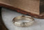 A close up image of The Elder King Band by The North Way Studio. It is a hand-carved golden wedding band, shown here in front of antique books atop a white marble tabletop.