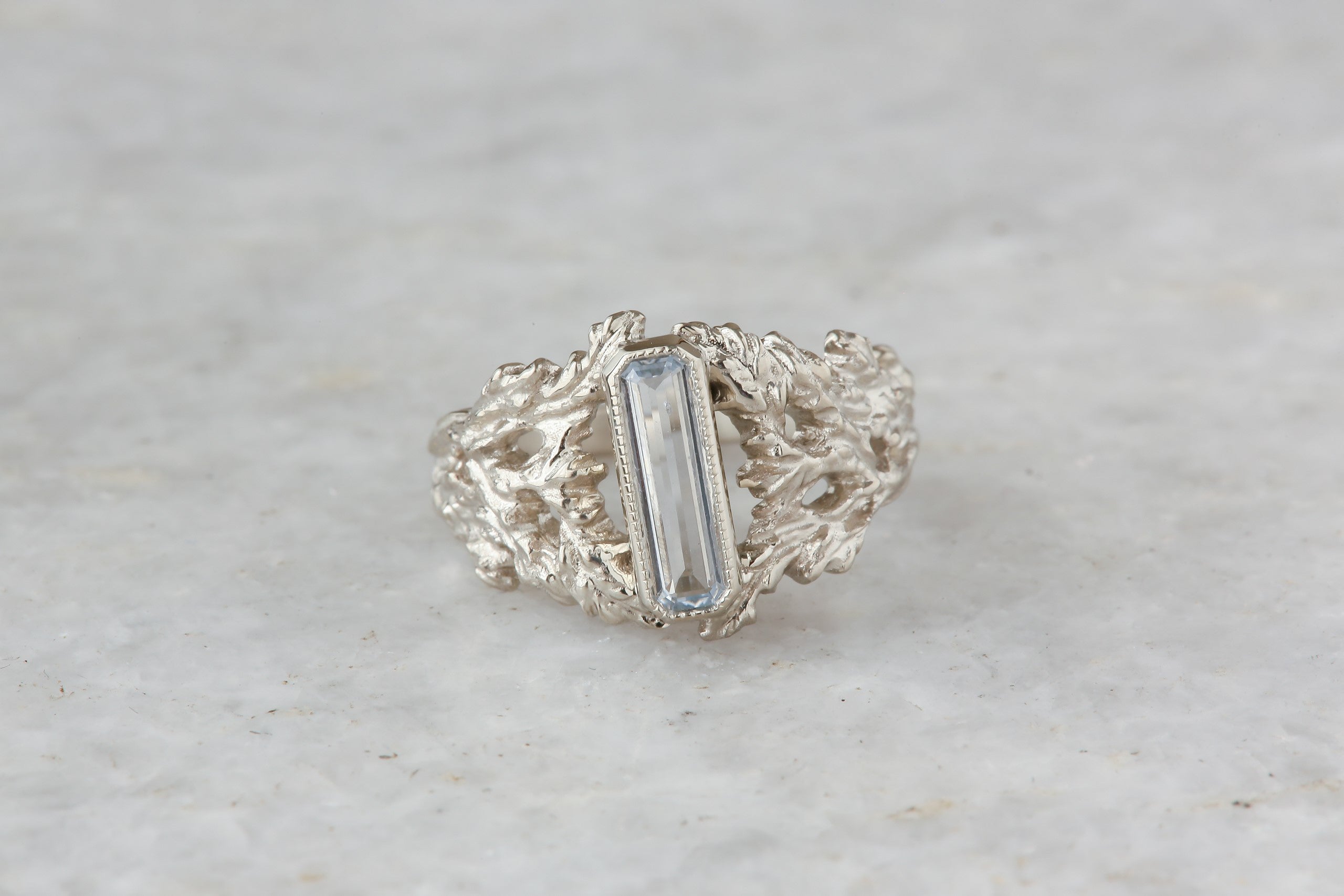 An elongated pale blue sapphire sits within a hand-carved band of white gold in this ring designed by The North Way Studio. The ready to ship engagement ring is shown in a closeup. 