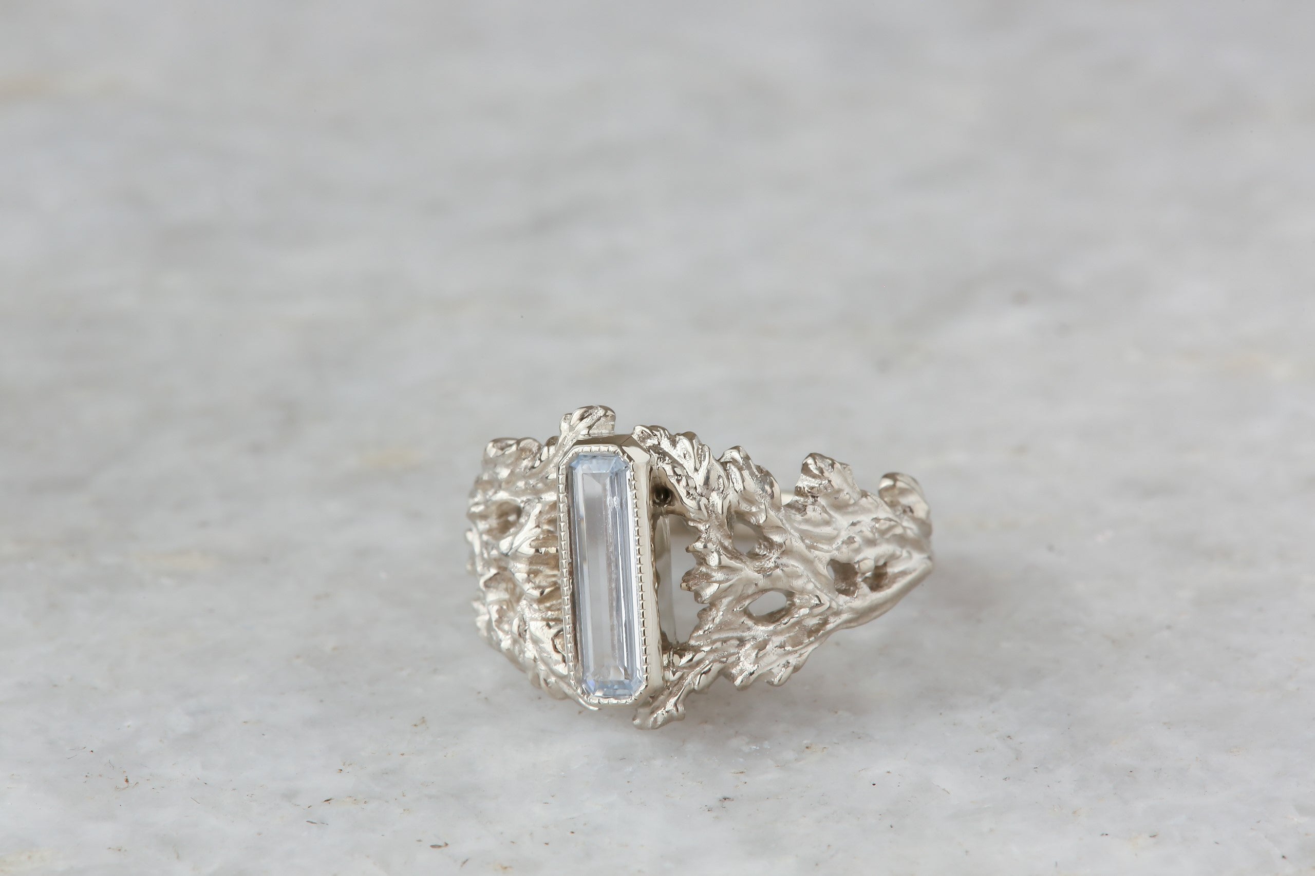 An elongated pale blue sapphire sits within a hand-carved band of white gold in this ring designed by The North Way Studio. The ready to ship engagement ring is shown in a closeup facing slightly left.