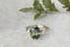 A fantasy engagement ring featuring a dark forest green natural oval brilliant cut sapphire, two marquise diamonds, and hand-carved leaf and bark texture in reclaimed 18 karat white gold. This fairytale garden engagement ring is ready to ship, size 6 1/2. Crafted by The North Way Studio in collaboration with Claire Pettibone.