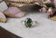 A dark forest green natural oval brilliant cut sapphire with diamonds engagement ring. Reclaimed 18 karat white gold with hand carved leaf band. This fairy engagement ring is shown resting on a marble surface beside dried roses and leaves.