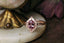 This custom engagement ring setting crafted by The North Way Studio features unfurling, carved leaves placed amongst a golden shield and a unique center stone of choice. This engagement ring is shown here in reclaimed 14 karat rose gold with a pink garnet center resting on an antique facing the viewer's left.