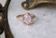 This custom engagement ring setting crafted by The North Way Studio features unfurling, carved leaves placed amongst a golden shield and a unique center stone of choice. This engagement ring is shown here in reclaimed 14 karat rose gold with an opalescent pink sapphire center resting on a marble surface beside an antique gold jewelry box.