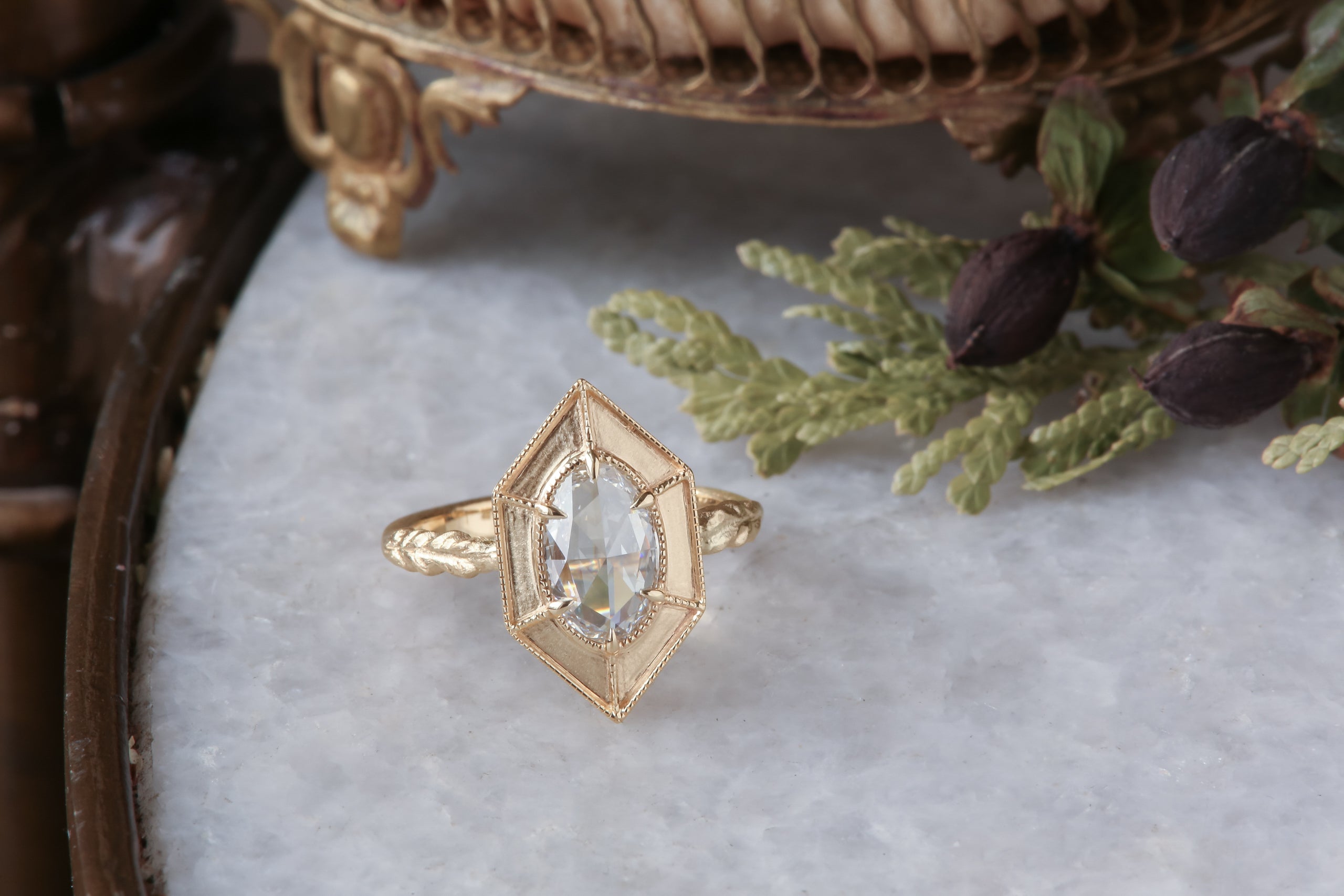 A relic of whittled gold and hues of an awakening gilded dawn. Carved 14 karat yellow gold cocoons about an elongated oval rose cut lab grown diamond. The Helm Ring in lab grown oval rose cut diamond is a ready to ship ring designed and sold at The North Way Studio. This diamond ring is shown resting on a marble surface.