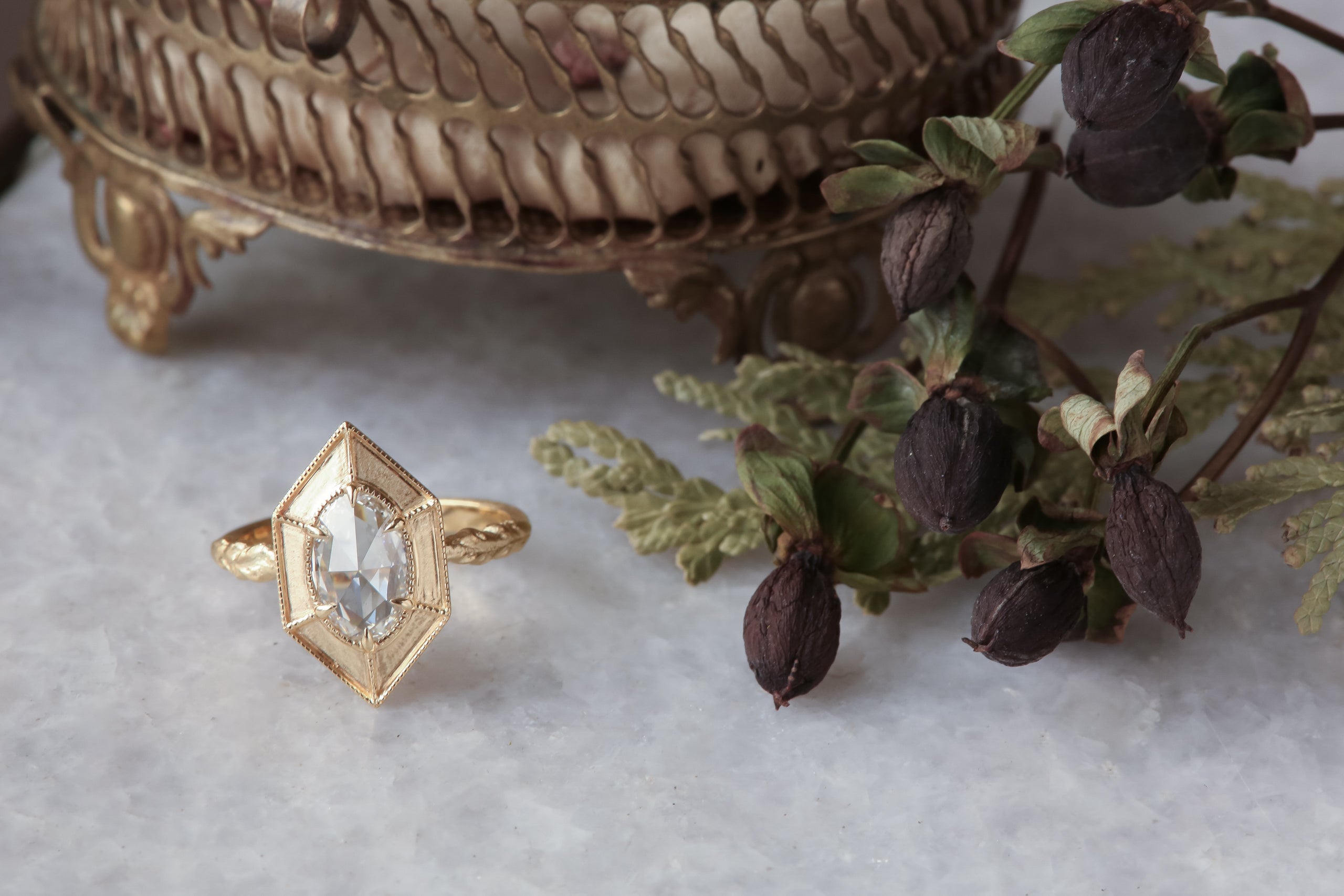 A relic of whittled gold and hues of an awakening gilded dawn. Carved 14 karat yellow gold cocoons about an elongated oval rose cut lab grown diamond. The Helm Ring in lab grown oval rose cut diamond is a ready to ship ring designed and sold at The North Way Studio. This diamond ring is shown facing the viewer on a marble surface.