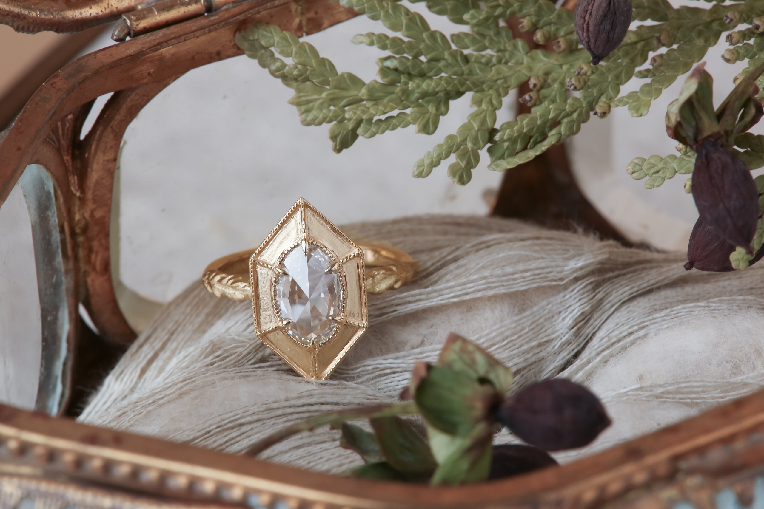 A relic of whittled gold and hues of an awakening gilded dawn. Carved 14 karat yellow gold cocoons about an elongated oval rose cut lab grown diamond. The Helm Ring in lab grown oval rose cut diamond is a ready to ship ring designed and sold at The North Way Studio. This fantasy engagement ring is shown facing the viewer and resting in an antique jewelry box.