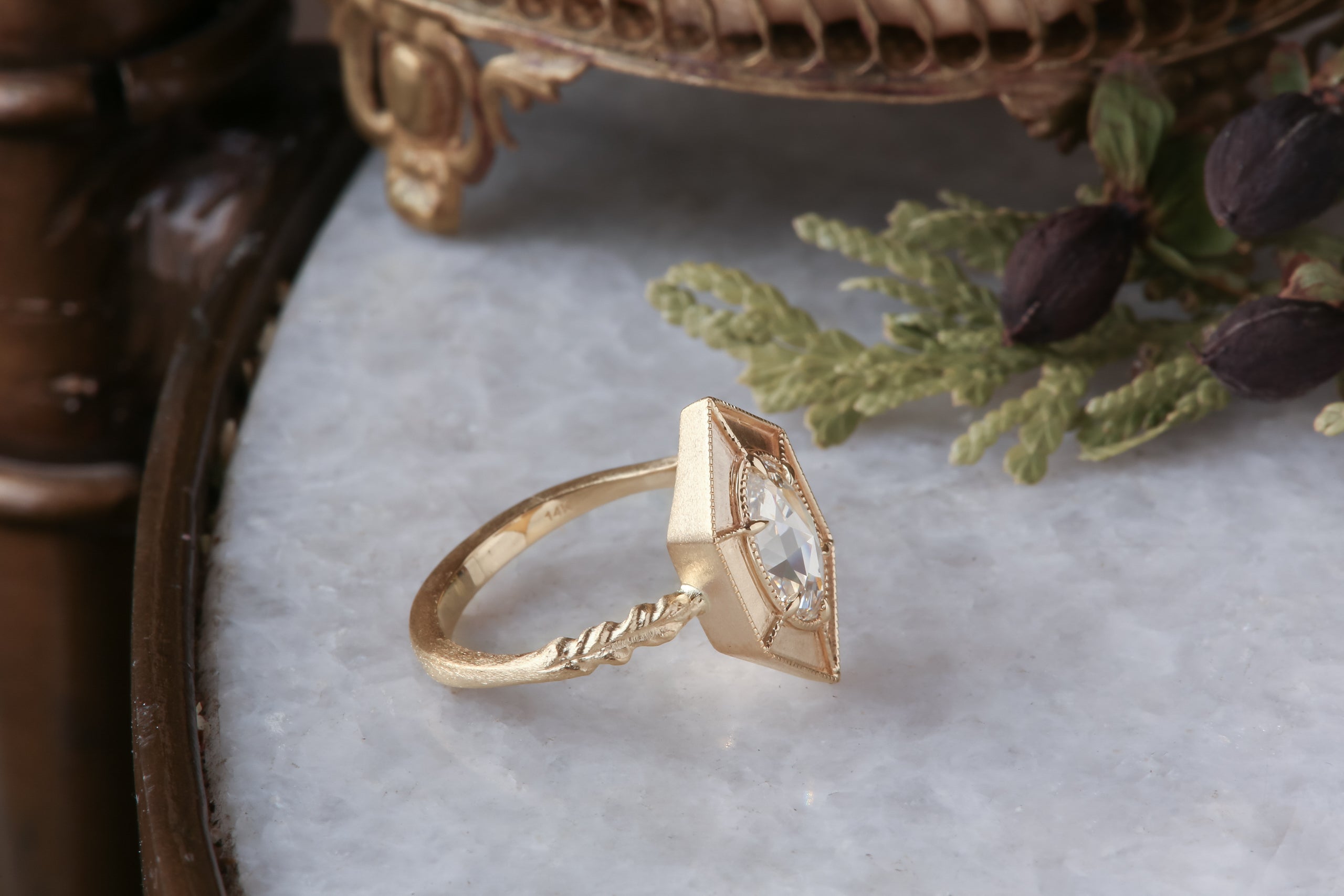 A relic of whittled gold and hues of an awakening gilded dawn. Carved 14 karat yellow gold cocoons about an elongated oval rose cut lab grown diamond. The Helm Ring in lab grown oval rose cut diamond is a ready to ship ring designed and sold at The North Way Studio. This fantasy engagement ring is shown resting on a marble surface.