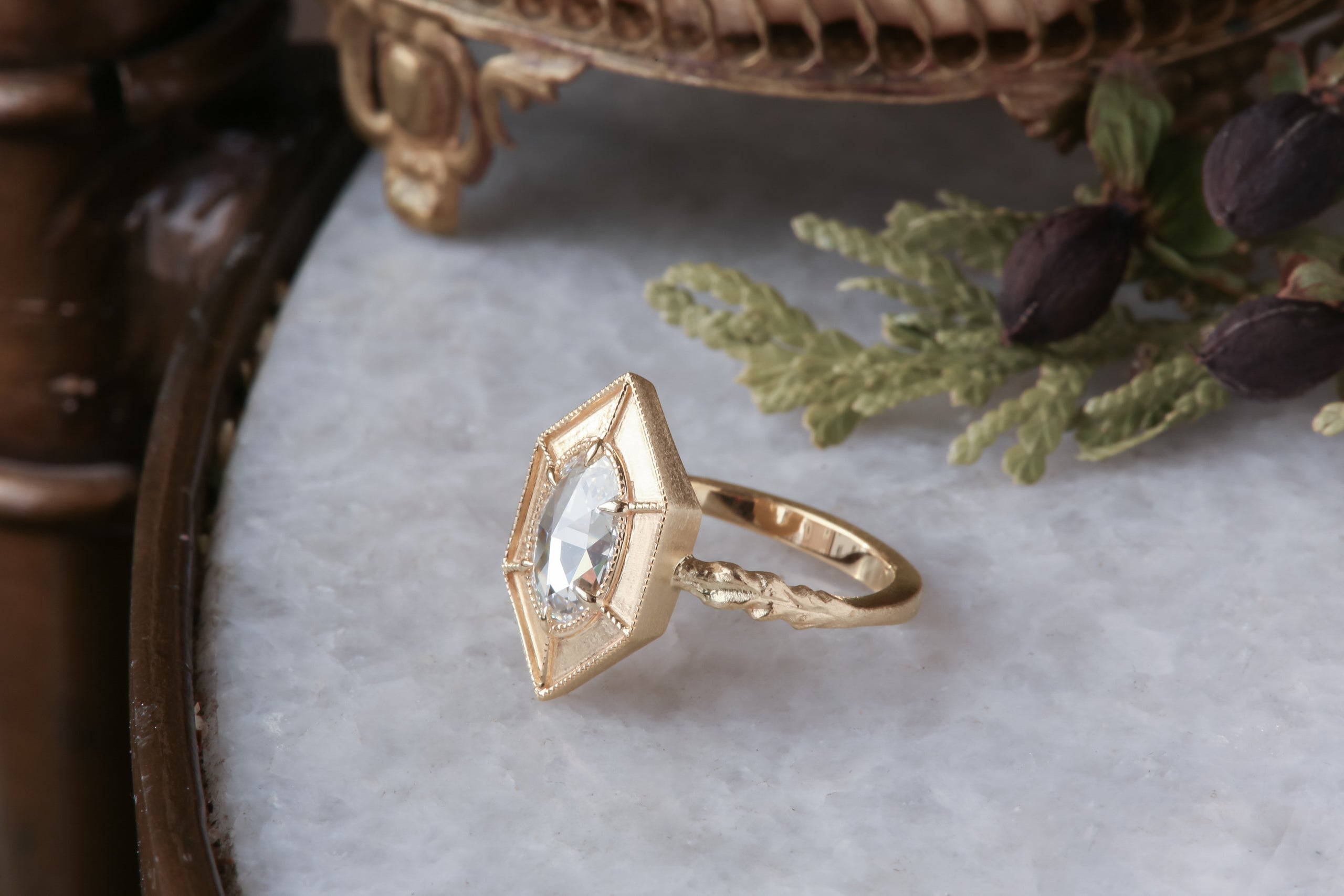 A relic of whittled gold and hues of an awakening gilded dawn. Carved 14 karat yellow gold cocoons about an elongated oval rose cut lab grown diamond. The Helm Ring in lab grown oval rose cut diamond is a ready to ship ring designed and sold at The North Way Studio. This diamond ring is shown resting on a marble surface.