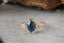 A large teal blue sapphire sits in the middle of this engagement ring setting designed by The North Way Studio shown sitting on a marble surface. A halo of diamonds surrounds the center stone and whittled bark and leaves are carved into the 14 karat gold band.