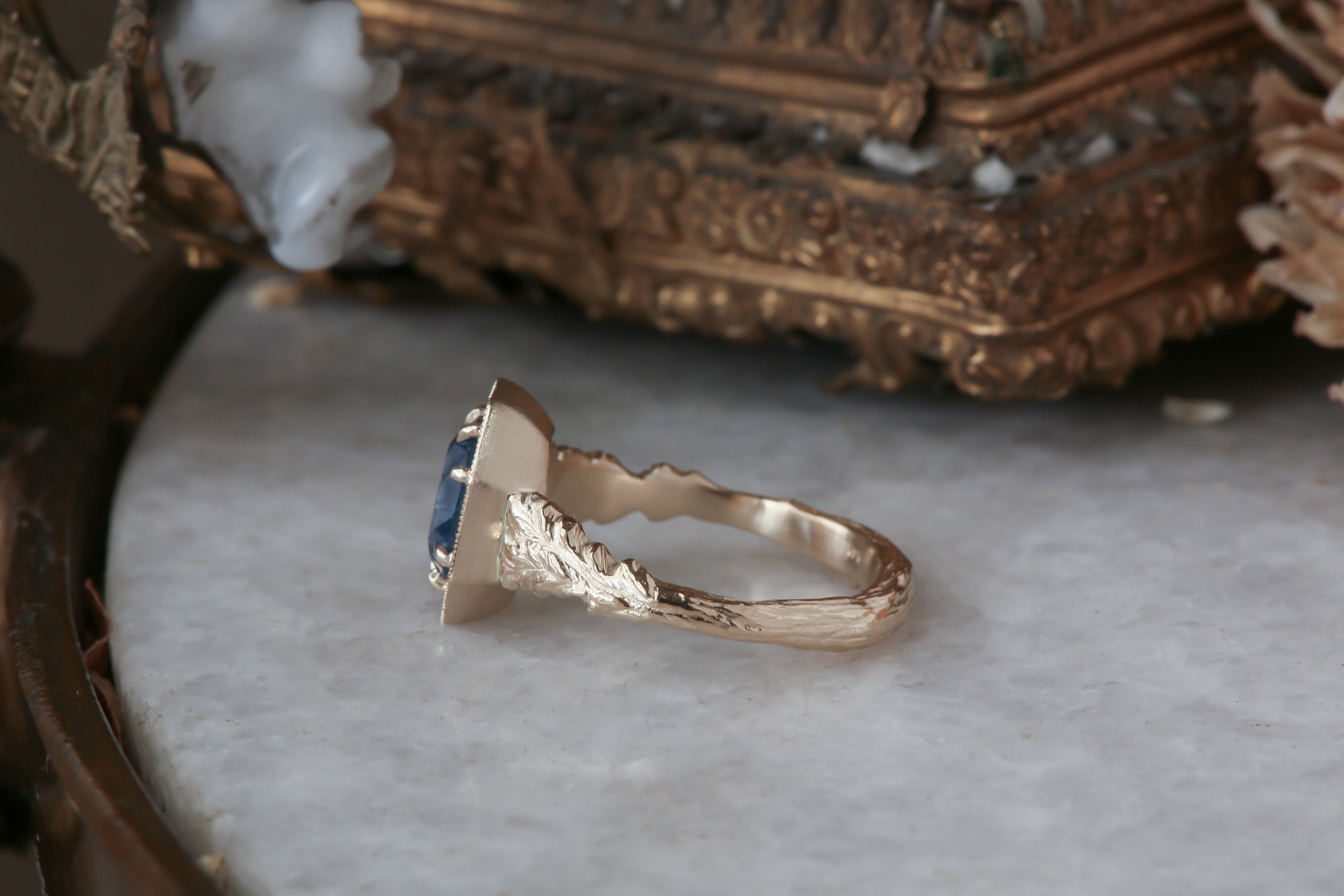 A large blue sapphire sits in the middle of this ready to ship engagement ring designed by The North Way Studio shown facing far to the left. A halo of diamonds surrounds the center stone. The band is hand-carved reclaimed gold.