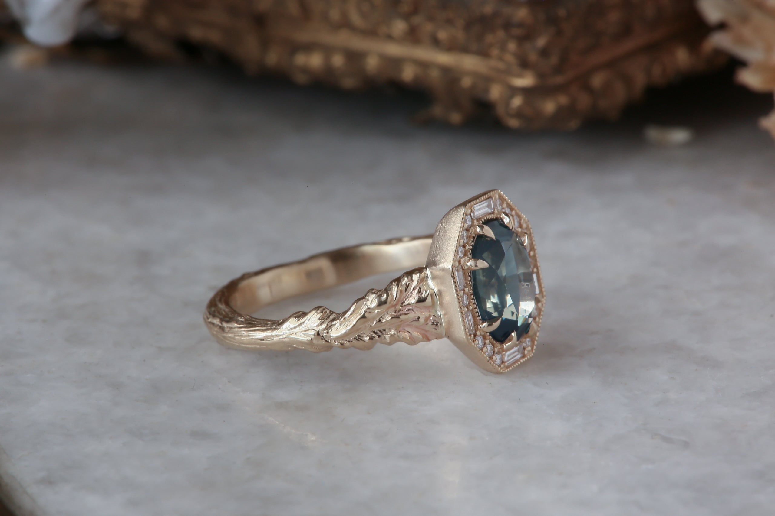 A large blue sapphire sits in the middle of this ready to ship engagement ring designed by The North Way Studio shown facing far right. A halo of diamonds surrounds the center stone. The band is hand-carved reclaimed gold.