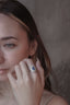 A blue sapphire engagement ring with a diamond halo designed by The North Way Studio in collaboration with Claire Pettibone and cast in reclaimed gold is shown on the hand of a young woman.