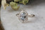 This heirloom engagement ring setting crafted by The North Way Studio features a shield halo of diamonds, engraved walls, and hand-carved leaves all strewn about a unique center stone of choice. This ring is shown in 14 karat white gold with a natural pale gray spinel center stone facing the viewer while resting on a marble table beside dried leaves.