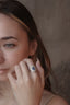 A blue sapphire ring with a diamond halo designed and created by The North Way Studio in collaboration with Claire Pettibone and cast in reclaimed gold is shown upon the hand of a young woman gently holding her hand to her face.