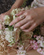 An engagement ring with natural pink spinel center, diamond halo, and sapphire leaves designed and crafted by The North Way Studio in collaboration with Claire Pettibone and cast in reclaimed 18 karat yellow gold is shown upon a hand resting on a bouquet of flowers.