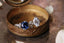 Reclaimed golden leaves are spun around your choice of center gemstone along with tiny diamond dew drops in this heirloom engagement ring setting designed and crafted by The North Way Studio. Two variations of this ring are shown resting in an antique jewelry box, both in 14 karat white gold with one featuring a round blue sapphire center and the other featuring an oval white moissanite center.