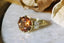 This heirloom engagement ring setting designed and crafted by The North Way Studio features two crescent diamonds and unfurling leaves of gold spun about a uniquely chosen center gemstone. This variation of The Sisters Three ring is shown cast in 18 karat green gold with a large round orange sapphire center. The ring is viewed close up as it rests facing slightly left on a marble table.