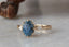 Marquise diamonds and unfurling carved gold leaves nestle a uniquely chosen center stone in this heirloom engagement ring setting designed and crafted by The North Way Studio. This ethereal engagement ring is shown cast in 14 karat yellow gold with a blue oval Montana sapphire center and resting on a marble surface. The ring is viewed close up providing a good look at the claw prongs.