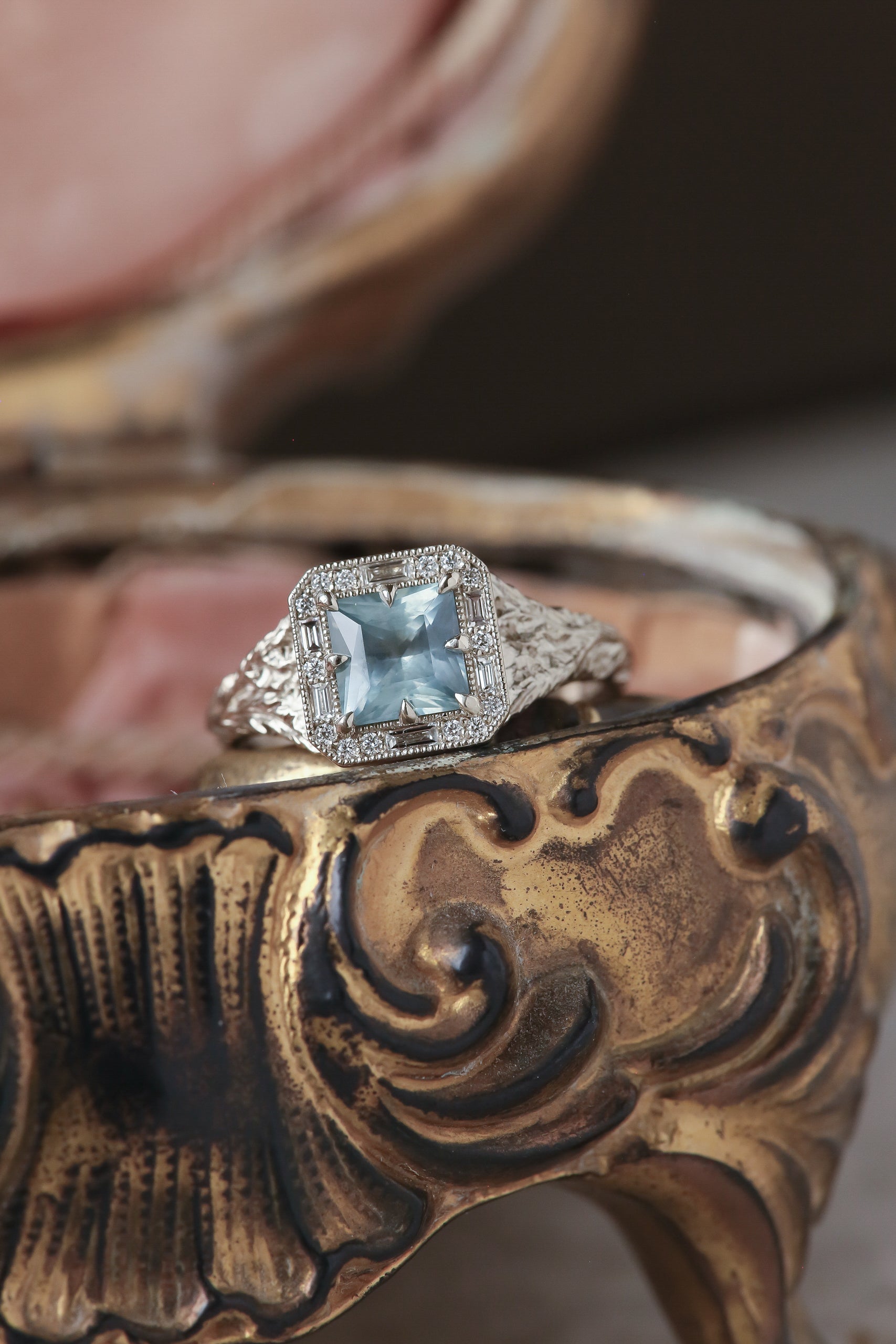 A square Montana sapphire sits within a halo of diamonds in this white gold, hand carved ring designed by The North Way Studio, shown in vertical atop a brass box.