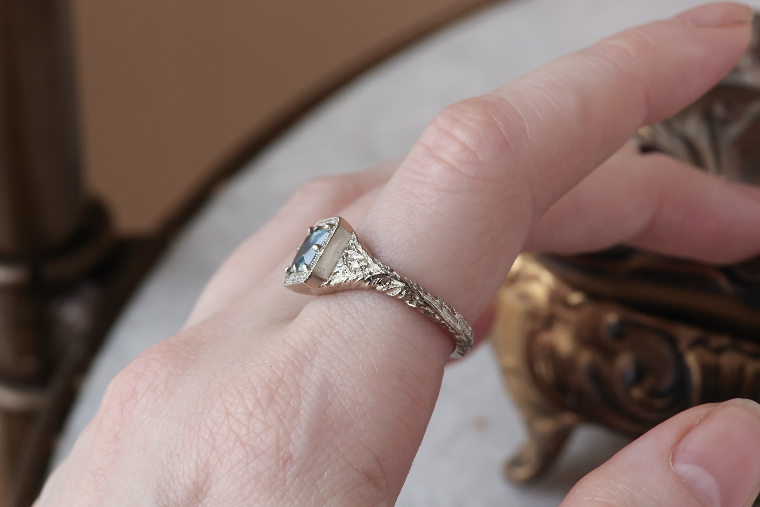 A princess cut Montana sapphire sits within a halo of diamonds in this white gold, hand carved ring designed by The North Way Studio. Shown worn on the first finger of a woman's hand.