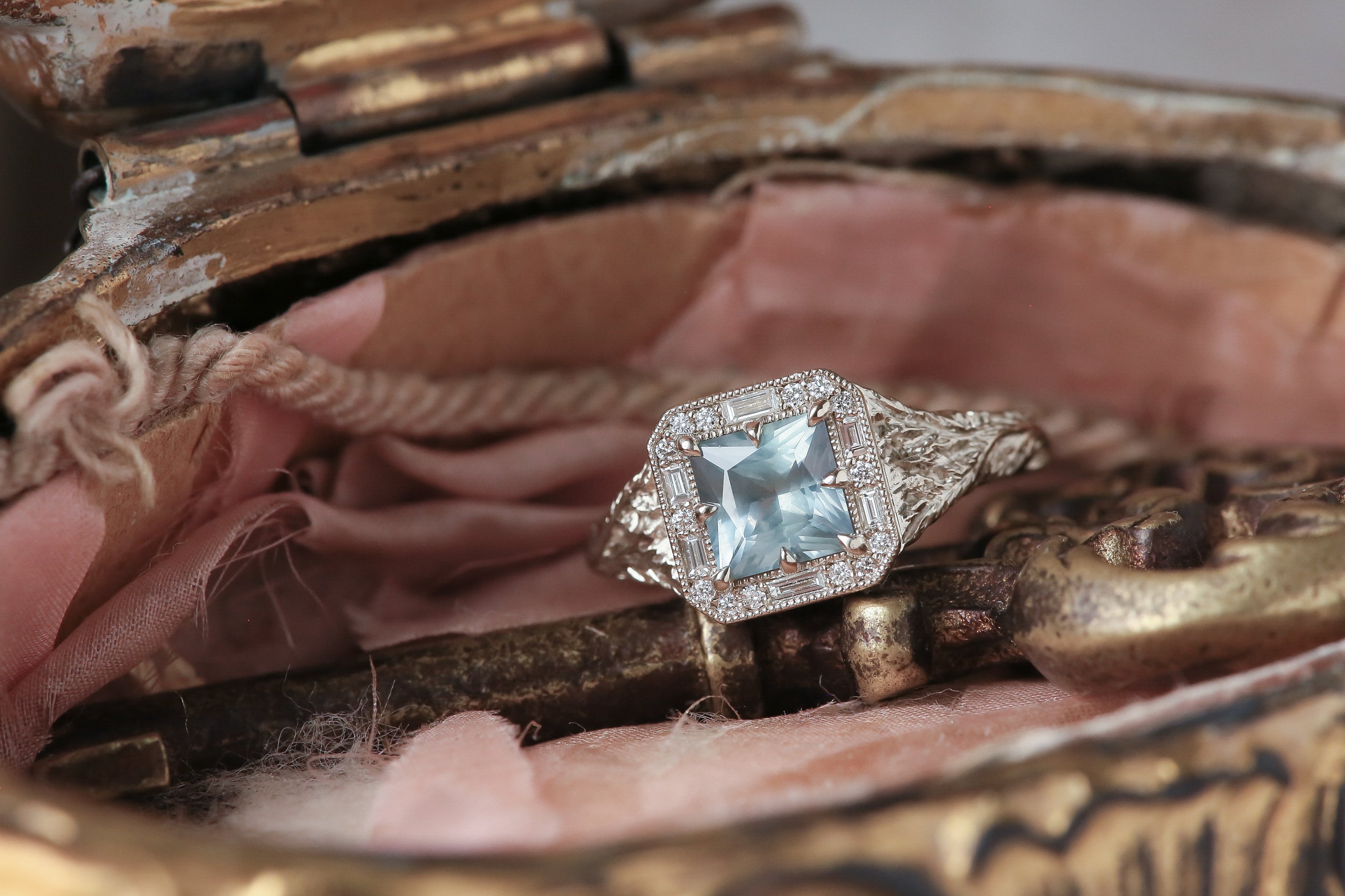 A square light blue silken Montana sapphire sits within a halo of diamonds in this white gold, hand carved ring designed by The North Way Studio. Shown within an antique jewelry box. 