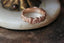 A golden wedding band designed by The North Way Studio. Hand-carved Scottish thistle and heather leaves shine in reclaimed gold. Shown atop a white marble tabletop.