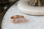 A golden wedding band designed by The North Way Studio. Hand-carved Scottish thistle and heather leaves shine in reclaimed gold. Shown from the side in a close-up, atop a white marble tabletop.