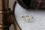 This heirloom engagement ring setting crafted by The North Way Studio features unfurling leaves and bark of carved gold, with petite claws to embrace a uniquely chosen center stone. This mythical ring is shown in 14 karat yellow gold with a pear cut diamond center. The engagement ring is shown resting on a marble table with an antique worn leather case in the background.