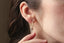 A small gold charm with lab grown pear diamond and hand-carved leaf details. Shown in white gold hanging on the Fae Hoop earrings on a woman's ear.