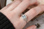 Fantasy elven alternative engagement ring designed and crafted by The North Way Studio. This ready to ship fairytale engagement ring features a diamond halo, a blue princess cut sapphire center, and hand-carved leaves and vines in reclaimed 18 karat white gold. Shown on a woman's hand.