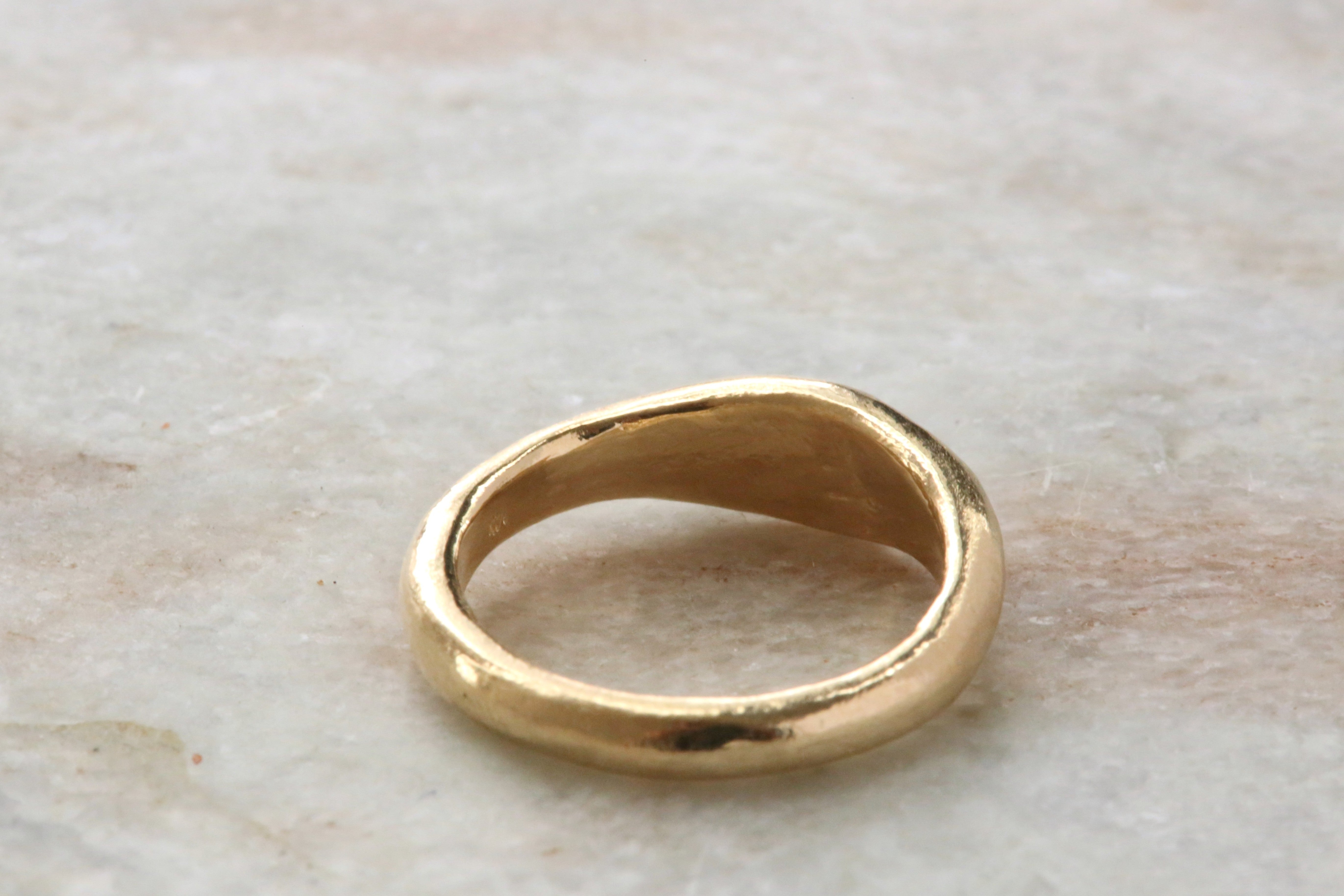 A gold signet-style ring with a thick, organic-shaped band, resting on a light beige marble surface.