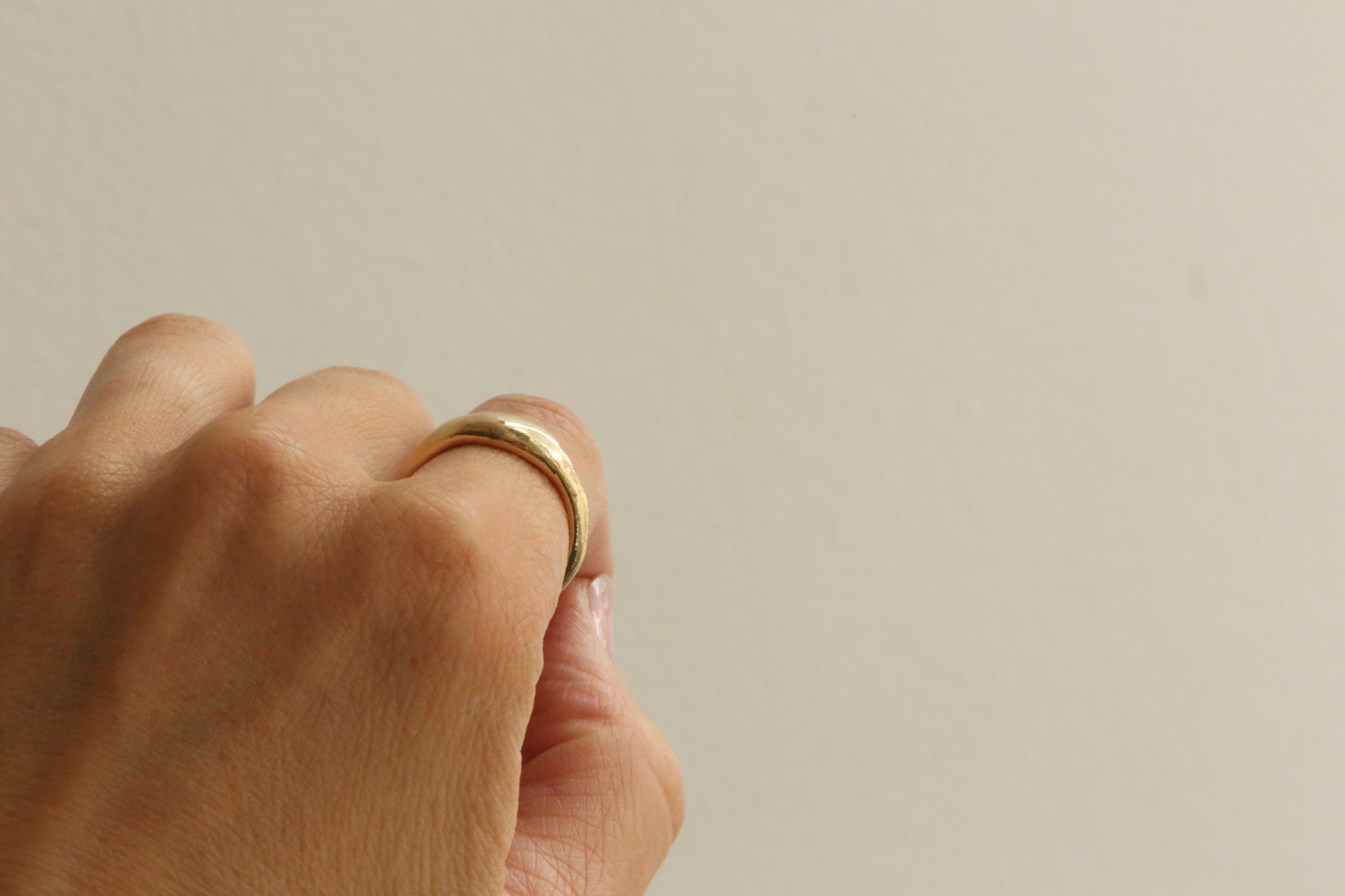 A close-up of a hand wearing a simple gold band ring against a plain light beige background.