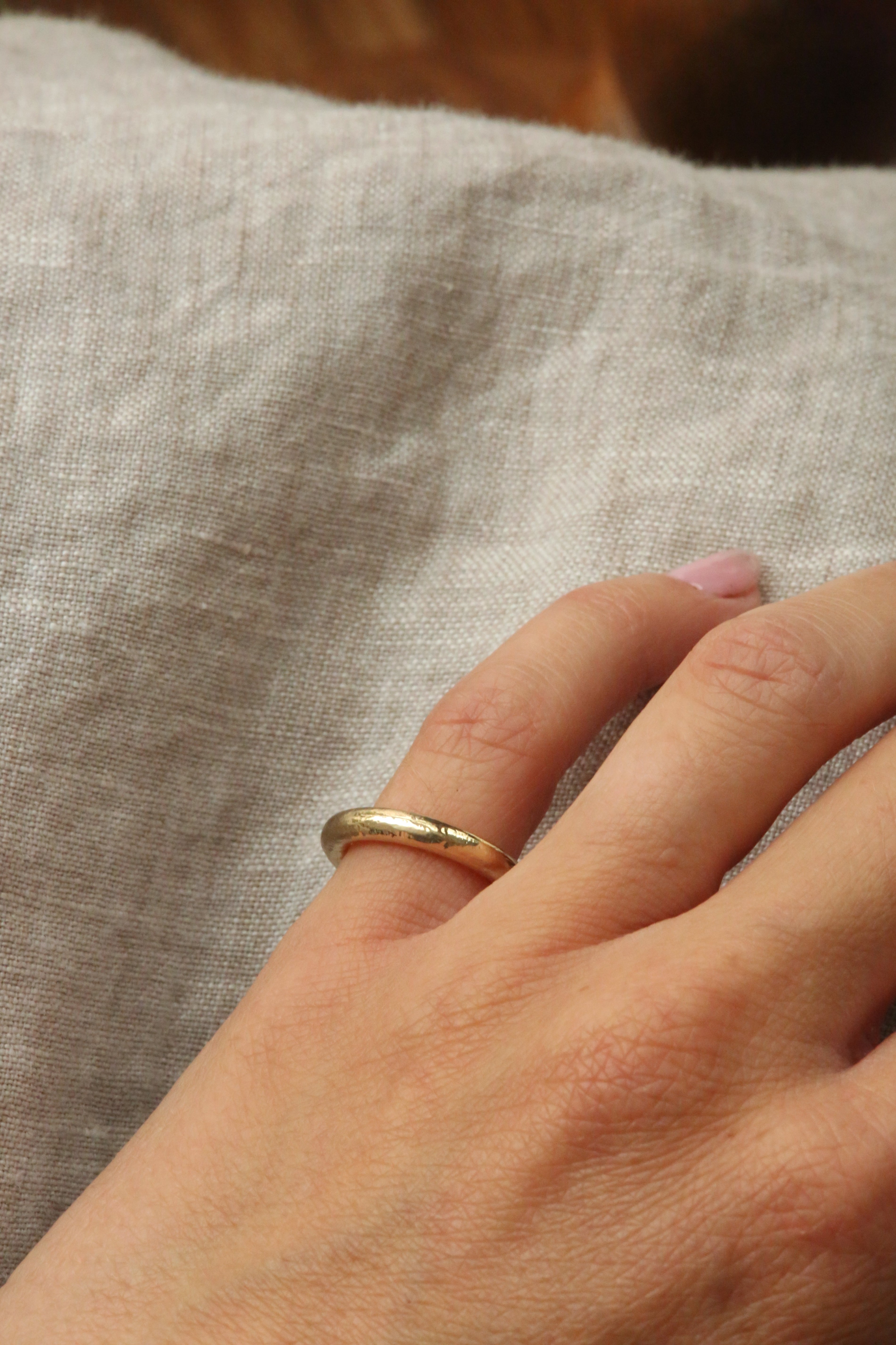 A close-up of a person's hand wearing a simple gold band ring, resting on a textured beige linen fabric.