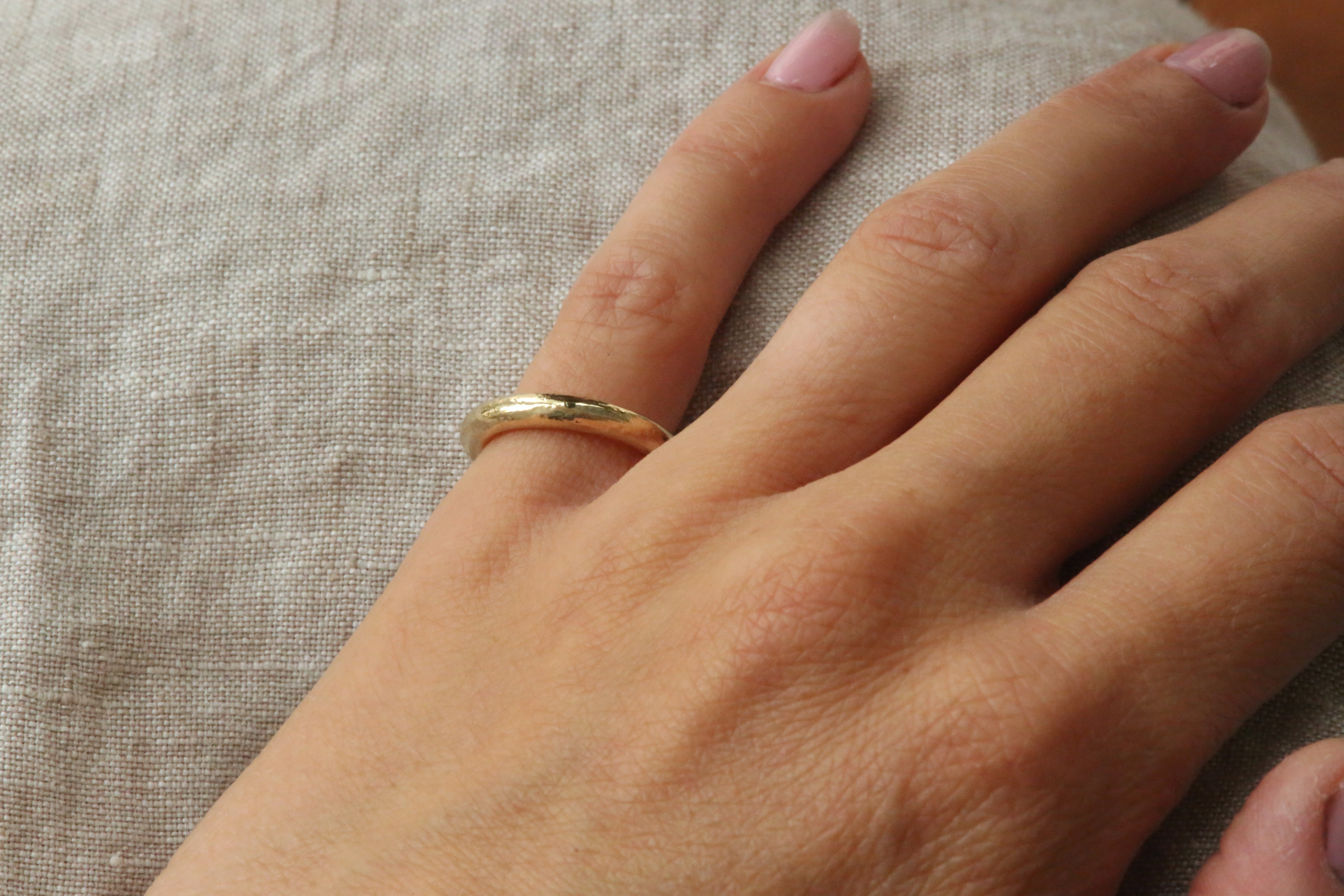 A close-up of a person's hand wearing a simple gold band ring on one finger, resting on a textured beige linen surface.