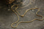 A gold snake chain necklace with a small gold clasp, laid out on a dark, textured metallic surface. Solid 14 karat yellow gold. 